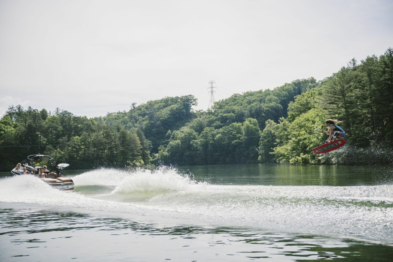 License-free image - A person is wakeboarding on a lake, performing a jump while being towed by a boat. The boat creates white water behind it. The scene is surrounded by lush green trees and a distant power line pylon. The sky is clear and bright.