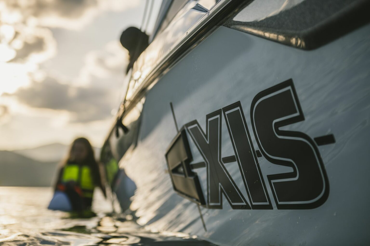 License-free image - Close-up of a boat titled &quot;AXIS&quot; on a calm body of water during sunset. Two individuals are in the water next to the boat, both wearing life jackets. The background features a partially cloudy sky with distant mountains.