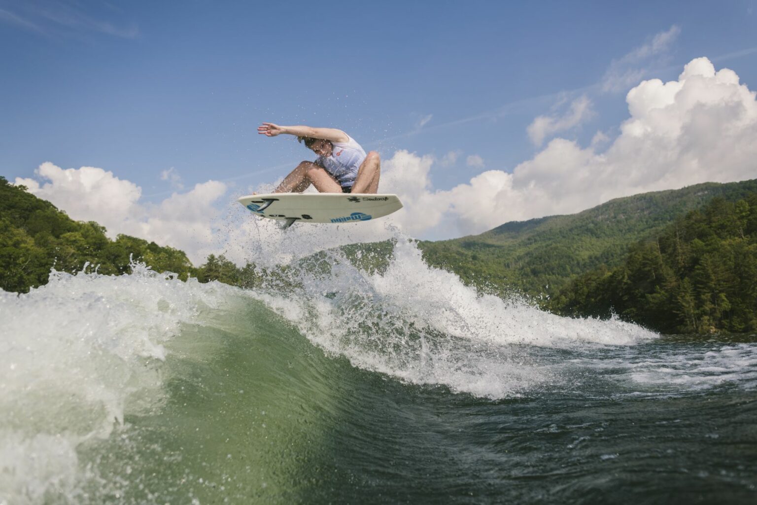 License-free image - A person is captured mid-air while wake surfing a large wave on a river surrounded by lush green hills and a partly cloudy sky. The water splashes around them as they perform a trick, showcasing their surfing skills.