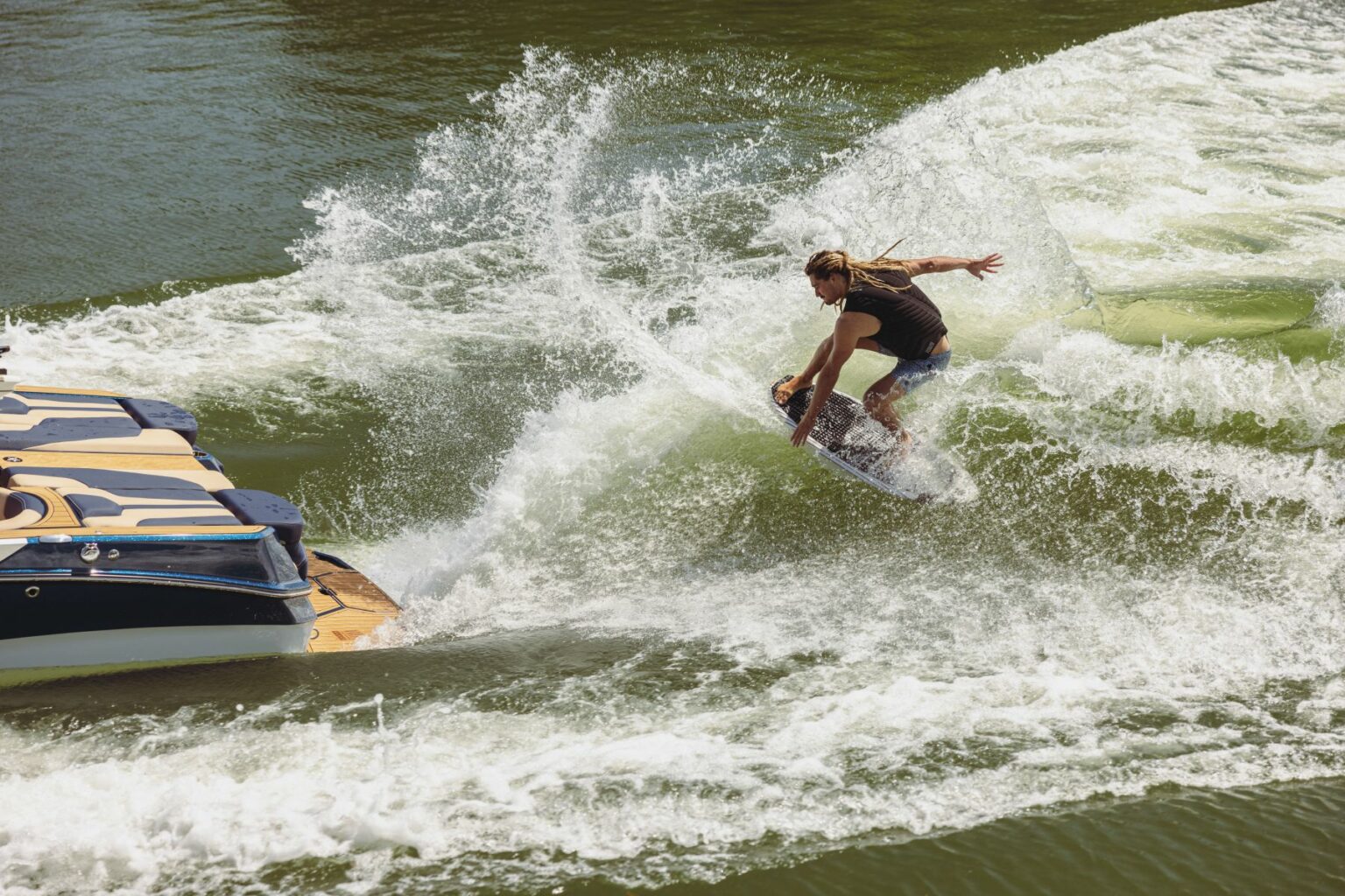 License-free image - A person rides a wakeboard behind a boat, performing a sharp turn on the water. Their body leans forward, kicking up a significant spray of water. The sun shines brightly, creating a vivid contrast between the white spray and greenish water.