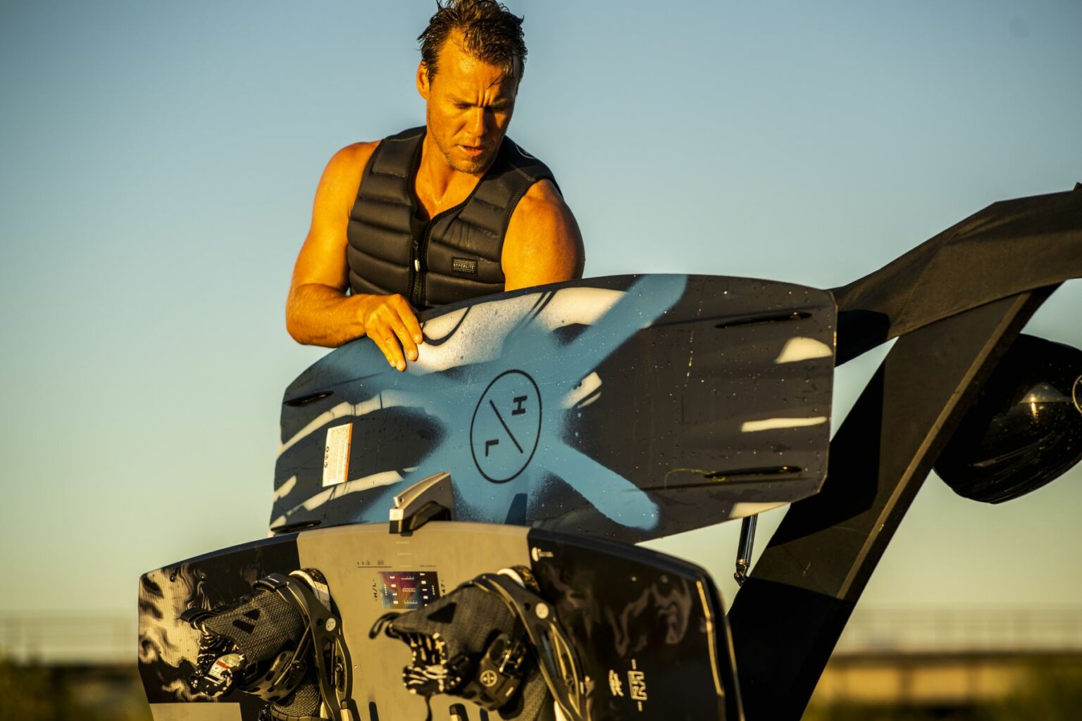 License-free image - A man wearing a black life vest is holding a wakeboard with blue and black patterns. He is attaching the wakeboard to a rack on a boat. The background shows a clear sky, suggesting it is a sunny day.