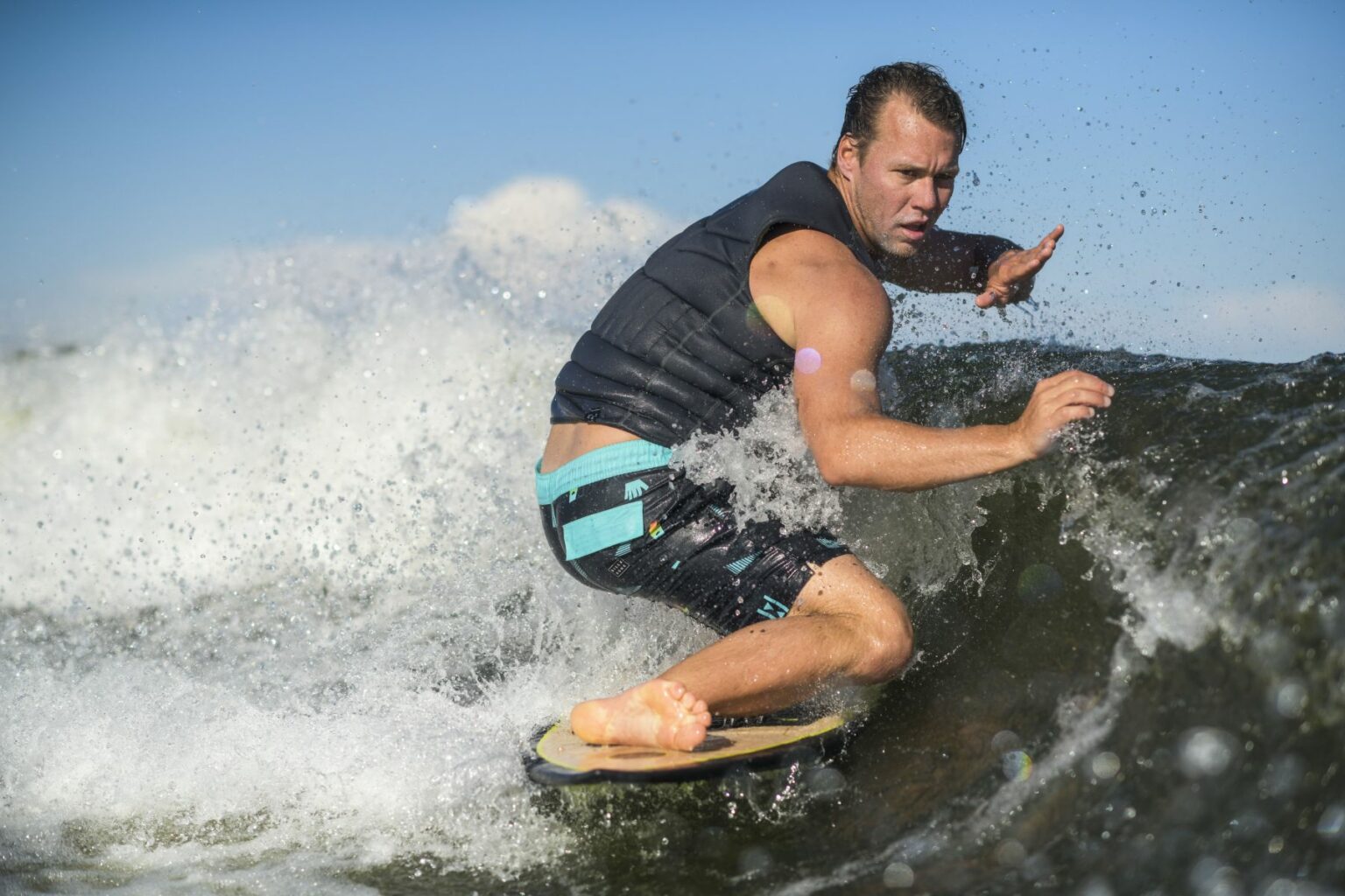 License-free image - A man rides a wave on a surfboard with a determined expression. He is wearing a sleeveless black life vest and blue-green board shorts. The sunny sky and ocean water surround him, with droplets of water splashing around as he navigates the wave.