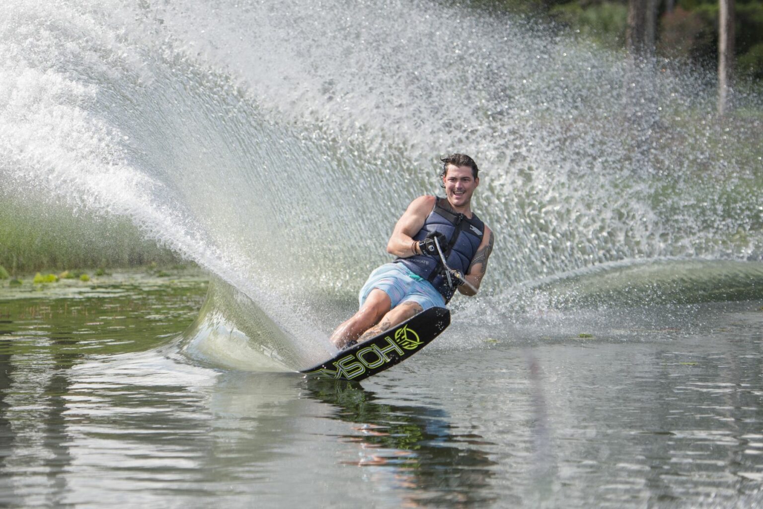 License-free image - A person in a life jacket and swim trunks is joyfully water skiing on a lake. They are holding onto the rope handle with both hands, and a large spray of water arcs behind them, indicating speed and skill. Trees and greenery can be seen in the background.