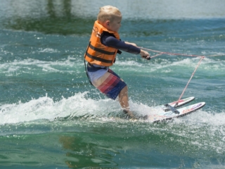 License-free image - A young boy wearing a safety vest water skiing on a sunny day. The child is holding onto a rope, balancing on two skis, and cutting through the wake with splashes of water around him. The water is clear with gentle waves, and the background is blurred.