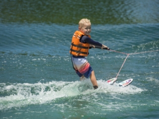 License-free image - A young child, wearing an orange life jacket and striped swim shorts, smiles while wakeboarding on a body of water. The child grasps the towrope handle and glides effortlessly over the water&#039;s surface, creating small waves in the wake.