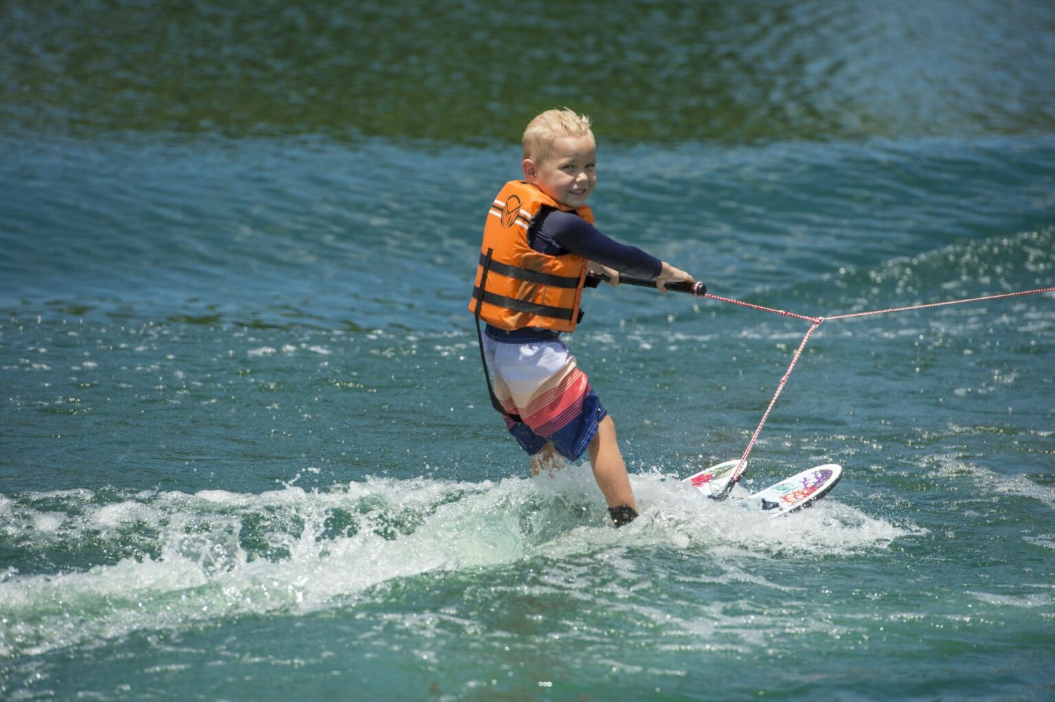 License-free image - A young child, wearing an orange life jacket and striped swim shorts, smiles while wakeboarding on a body of water. The child grasps the towrope handle and glides effortlessly over the water&#039;s surface, creating small waves in the wake.