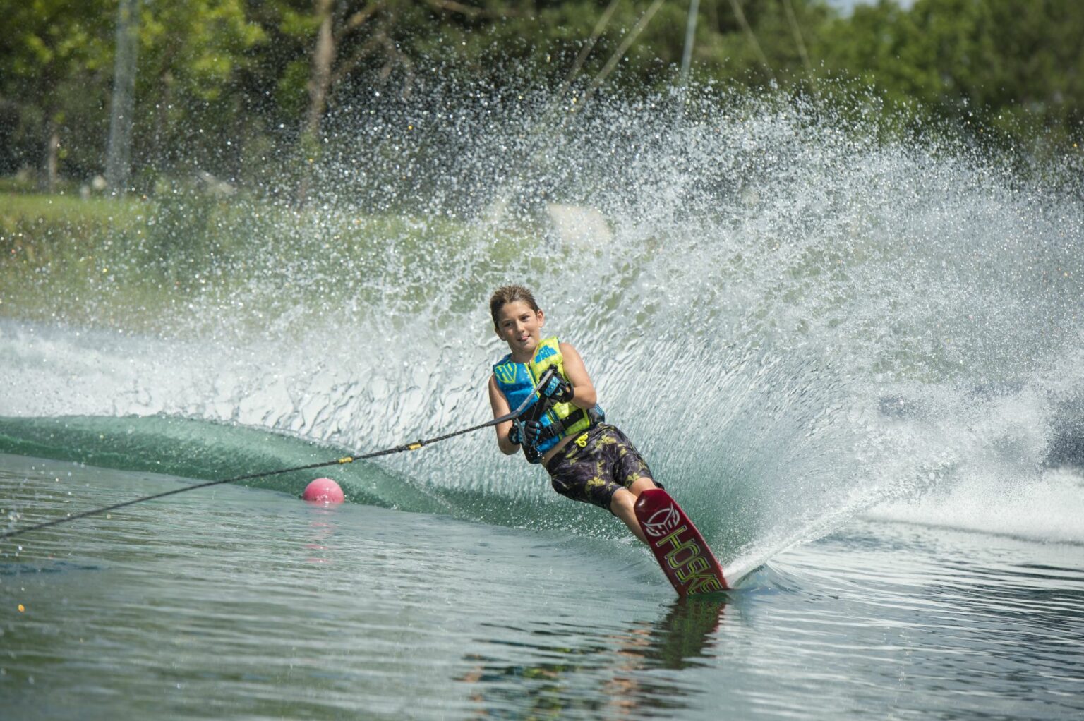 License-free image - A child is water skiing on a lake, creating a large spray of water behind them. They are leaning to one side with one hand holding onto the tow rope. The skier is wearing a blue life jacket and patterned shorts, with greenery visible in the background.