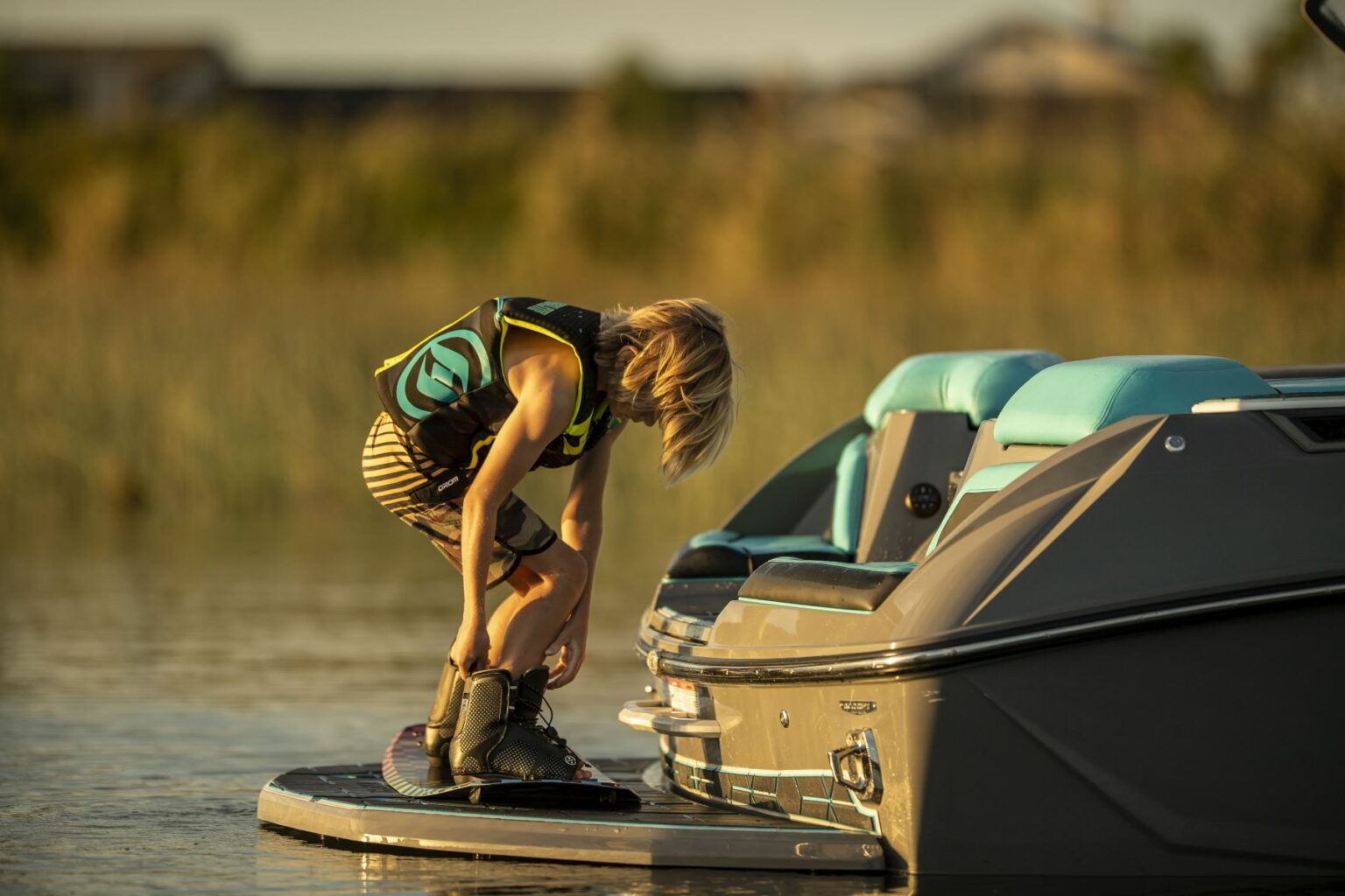 License-free image - A child wearing a life vest bends over to adjust their wakeboard bindings while standing on a wakeboard at the edge of a boat. The boat is in calm water with a natural, blurry background of reeds and buildings.