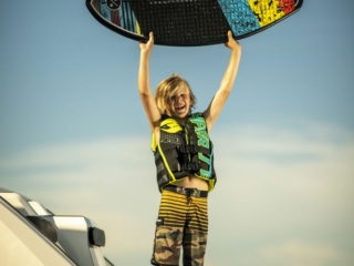License-free image - A child wearing a life jacket and striped shorts stands on a dock by a boat, holding a colorful wakeskate above their head triumphantly against a clear sky.