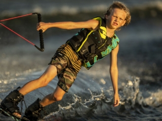 License-free image - A young child with blond hair rides a wakeboard on water, holding onto a rope with both hands. They are wearing a yellow and black life vest and camouflage-patterned shorts. The water splashes around them as they balance, with a determined expression on their face.