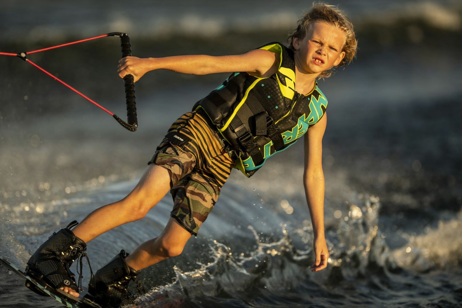 License-free image - A young child with blond hair rides a wakeboard on water, holding onto a rope with both hands. They are wearing a yellow and black life vest and camouflage-patterned shorts. The water splashes around them as they balance, with a determined expression on their face.