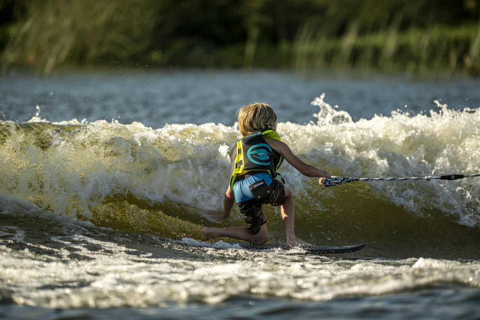 License-free image - A child with blonde hair, wearing a yellow life vest, is wake skating on a body of water. They are holding onto a tow rope and gliding over a small wave. The background shows greenery on the shore.