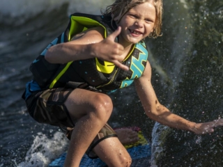 License-free image - A young person with wet blond hair, wearing a life vest, smiles and makes a shaka hand gesture while riding a wave on a surfboard behind a boat. The water splashes around them, and they appear to be enjoying the sunny day.
