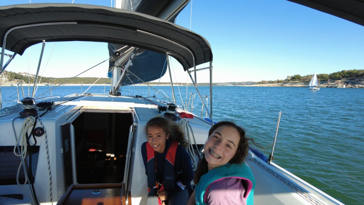 License-free image Two smiling girls on a sailboat in sunny weather. One is seated in the cockpit and the other is standing beside her. The boat is sailing on a calm body of water with blue skies and another sailboat visible in the background. Both girls are wearing life vests.