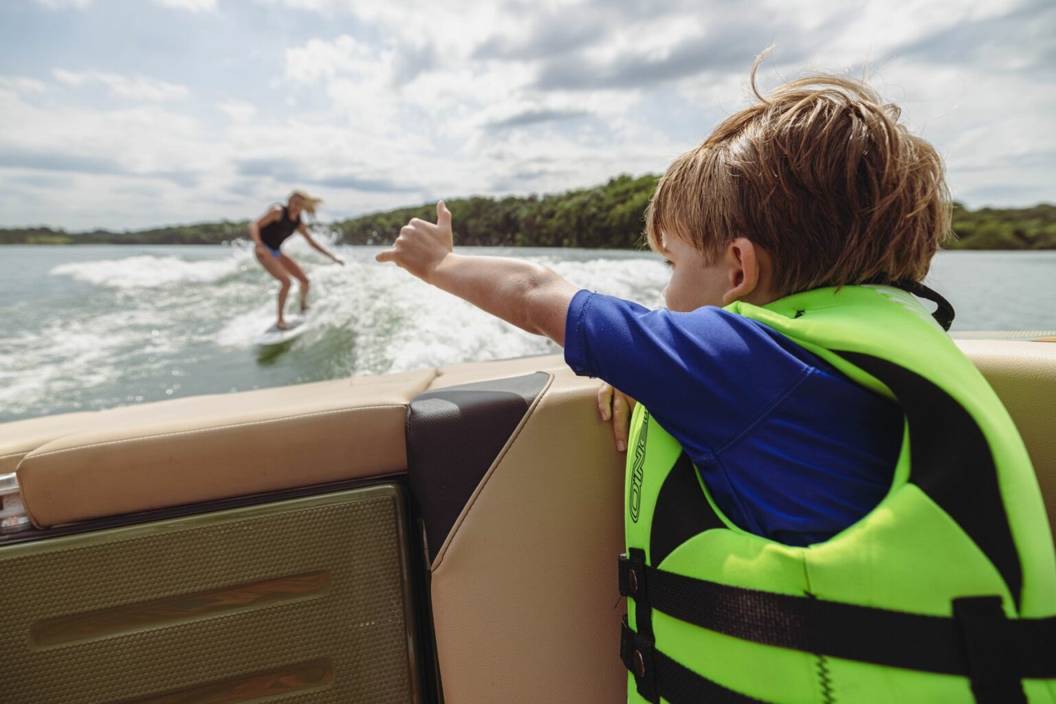 License-free image - A child wearing a bright green life jacket sits in a boat, giving a thumbs-up to a person wakeboarding on the water behind them. The wake surfer is riding a wave, and the background includes a cloudy sky and tree-lined shore.