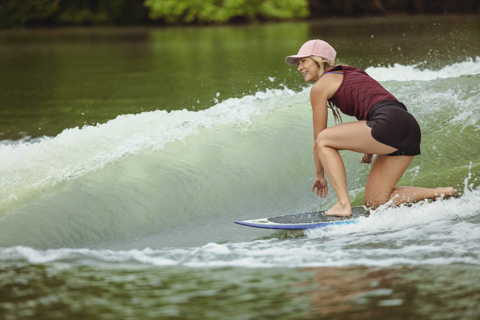 License-free image - A person wearing a pink cap, maroon tank top, and black shorts is surfing on a wave. The surfer is crouched low on the board, with a focused expression, surrounded by a lush green landscape and calm water.