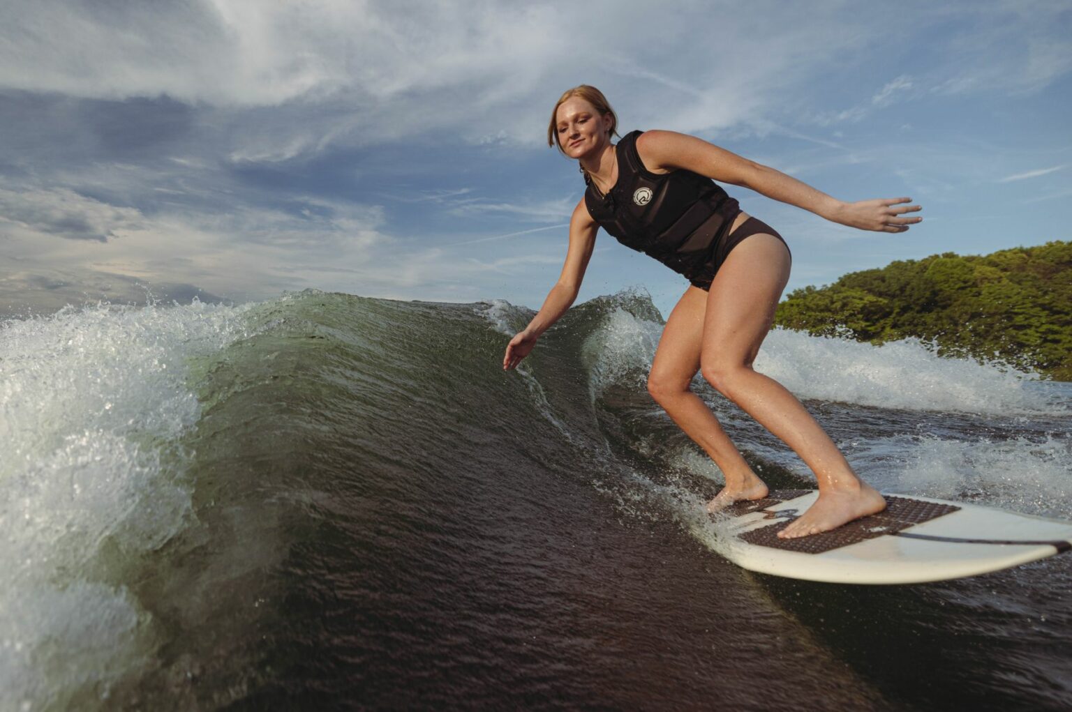 License-free image A person wearing a black life vest and black shorts is surfing on a wave. The sky is partly cloudy with patches of blue, and there is greenery visible in the background. The individual has a confident stance, balancing on a white surfboard.