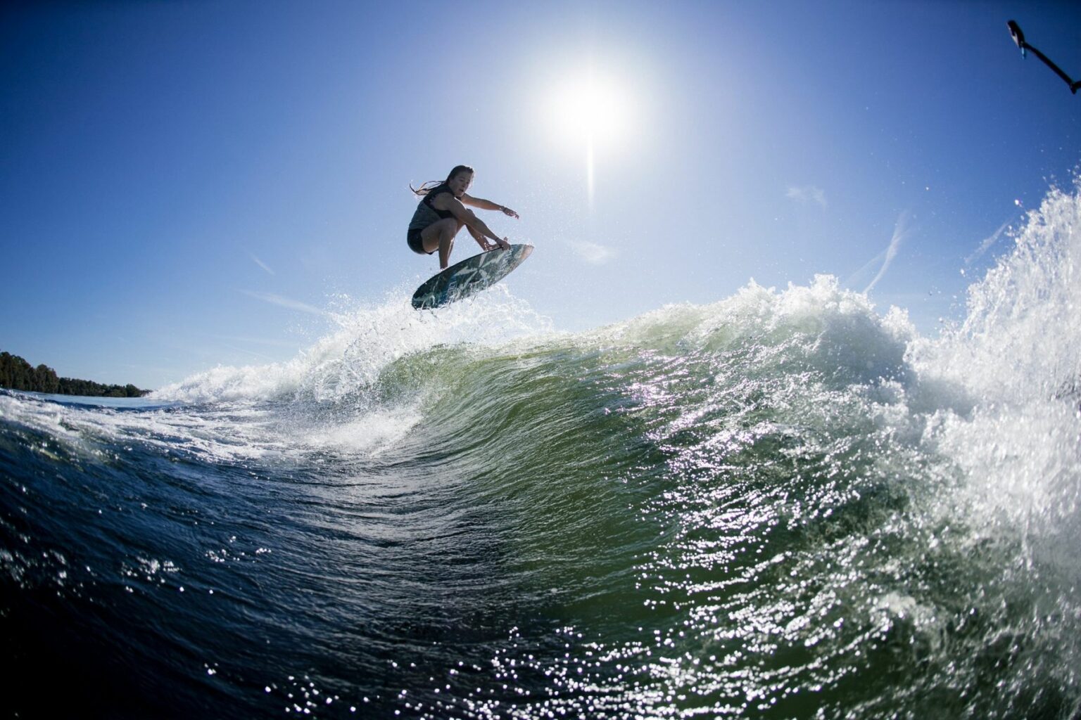 License-free image - A person surfing a large wave in bright sunlight, captured mid-air above the crest with clear blue skies and a shining sun overhead. The water is sparkling, and the wake surfer&#039;s silhouette is highlighted against the backdrop.