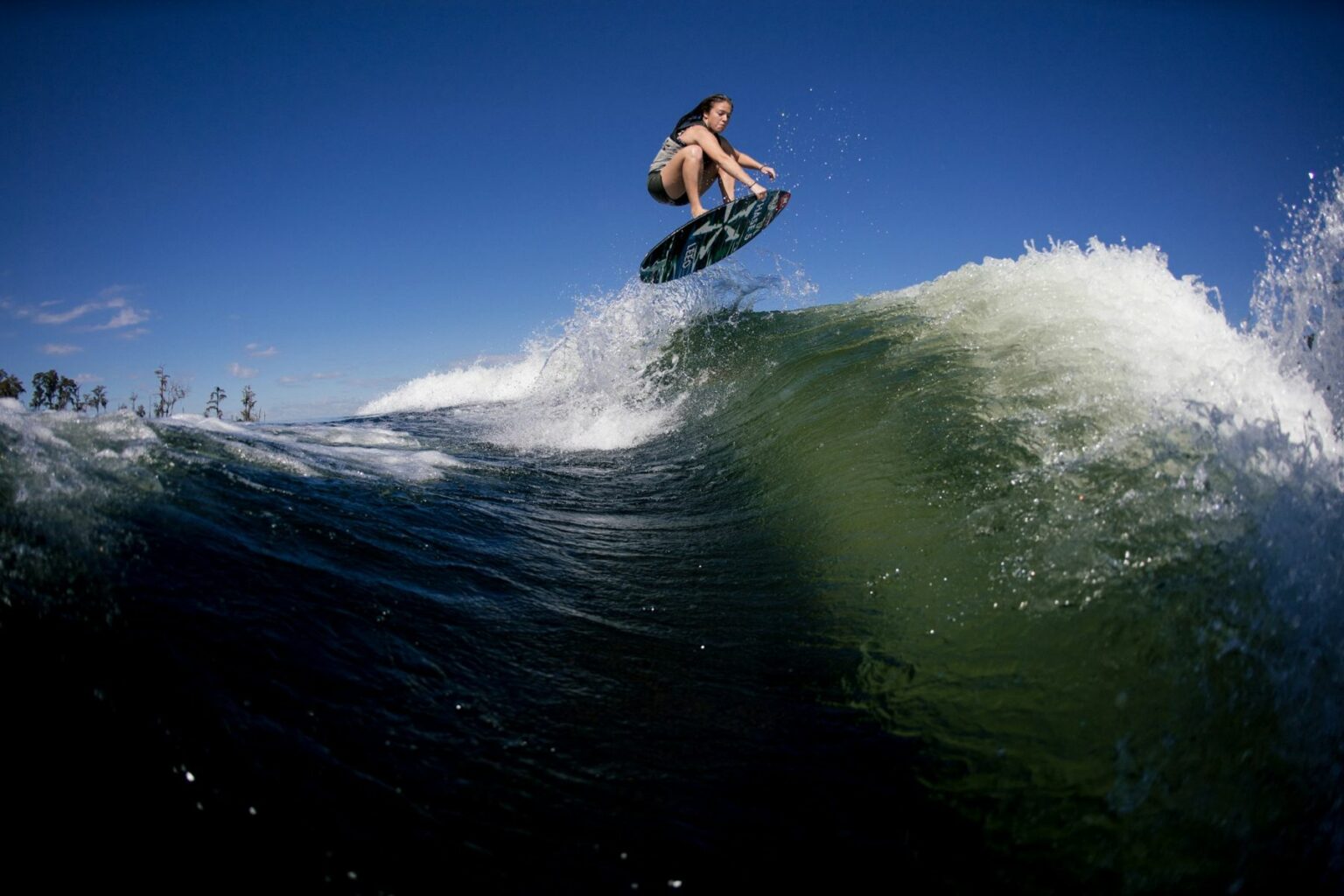 Royalty-free image - A person wearing swim shorts is performing an aerial trick on a wake surfboard, catching air above a large wave. The clear blue sky and white foam of the wave contrast with the deep green water below. Trees are faintly visible in the distant background.