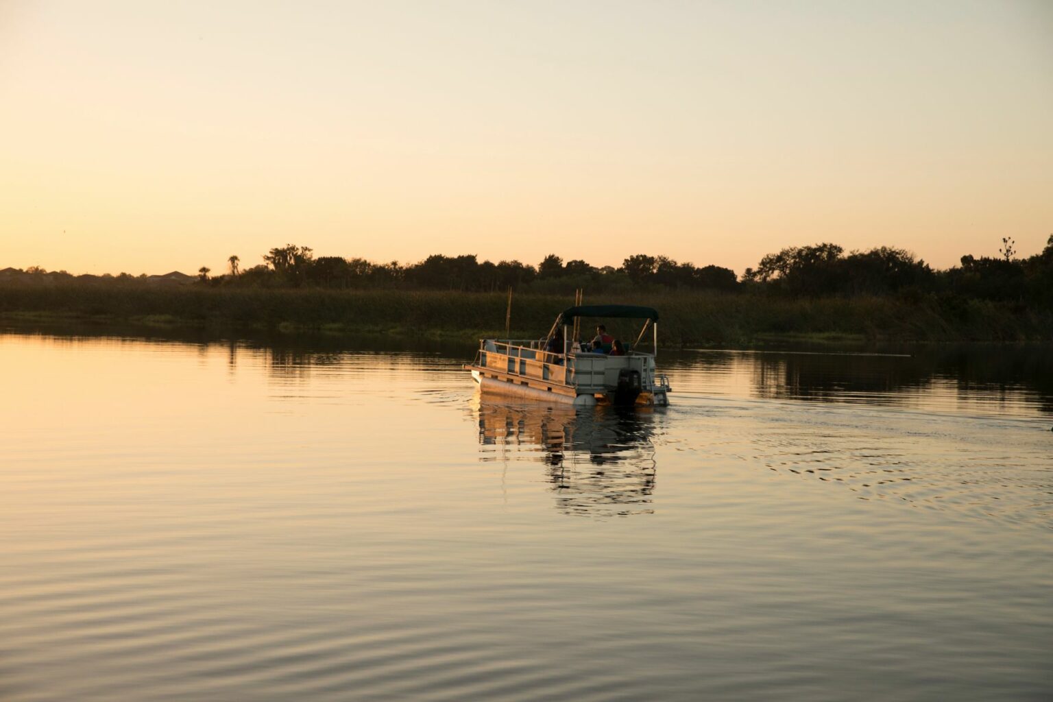 License-free image. A small pontoon boat navigates calm waters at sunset, leaving gentle ripples in its wake. The sky is a gradient of warm colors, and the horizon is lined with silhouetted trees and vegetation. The scene conveys tranquility and the beauty of nature.