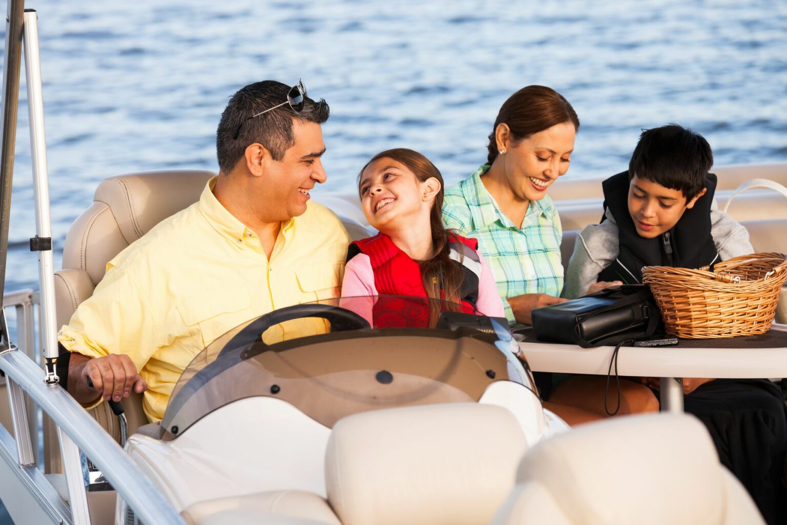 Royalty-free image - A family of four enjoys a boat ride. The father steers, wearing a yellow shirt, while the daughter sits beside him, smiling. The mother, in a green shirt, and the son, in a life jacket, sit at a table with a basket, overlooking the water.