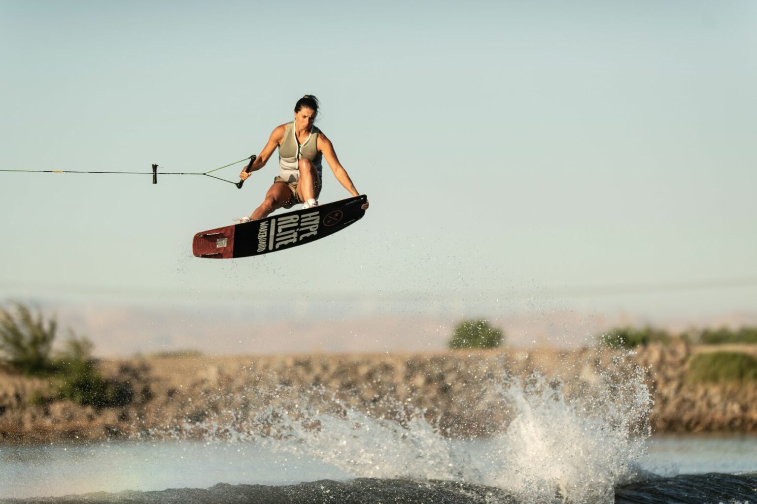 Royalty-free image - A person is wakeboarding, suspended in mid-air while holding onto a rope. They are wearing a grey life vest and the wakeboard features the text &quot;Hyperlite&quot; on the bottom. Water splashes below, and a distant shoreline is visible in the background.