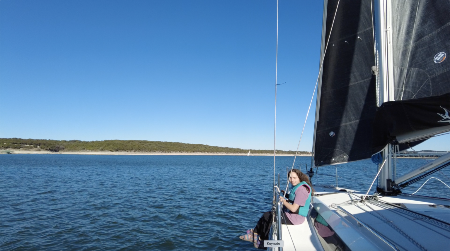 License-free image A person is sitting on the edge of a sailboat which is floating on calm, blue water. The person is wearing a life vest and appears relaxed. The sky is clear, and a coastline with sparse vegetation is visible in the background.