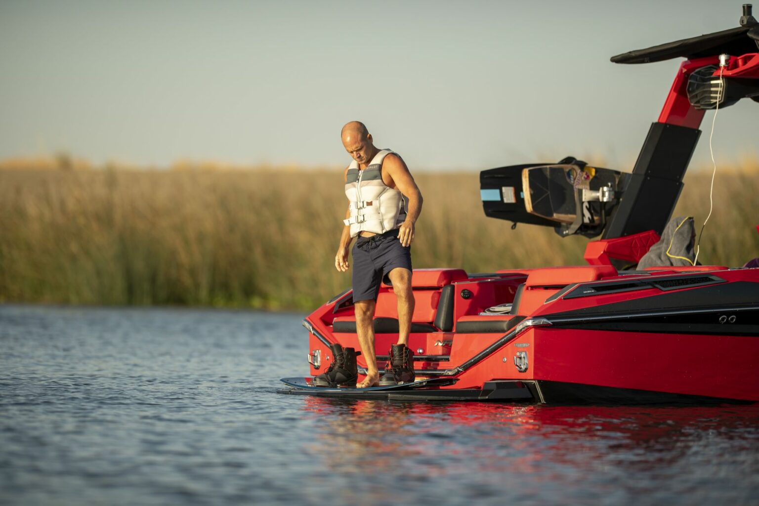 License-free image - A man in a life vest and shorts stands on a wakeboard attached to the back of a red wake boat on a calm body of water. He appears to be preparing to enter the water, with reeds visible in the background.