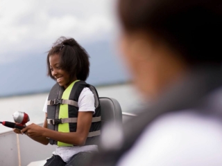 Royalty-free image A young girl, wearing a life jacket, smiles as she holds a fishing rod on a boat. Another person, blurred in the foreground, also wears a life jacket. The background features a calm body of water under a cloudy sky.