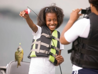 License-free image A young girl with an excited smile holds up a fishing line with a small fish she caught. She is wearing a life vest and is on a boat. Another person, also in a life vest, is taking her photo. The background is a blurred water scene.