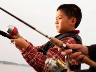 License-free image A young boy wearing a red plaid shirt and life vest concentrates while holding a fishing rod. Another person beside him, also holding a fishing rod, is visible but blurred in the foreground. The background shows a calm body of water under an overcast sky.
