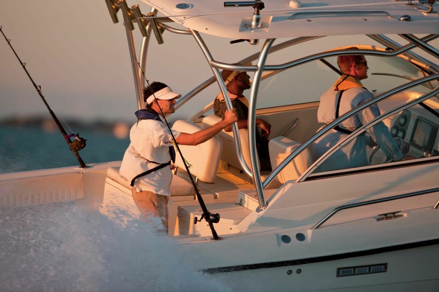 Royalty-free image. Two people are riding on a white motorboat during sunset. One person is standing, wearing a visor and holding a fishing rod, and the other is seated behind the wheel, steering the boat. The scene captures the dynamic motion of the boat cutting through the water.