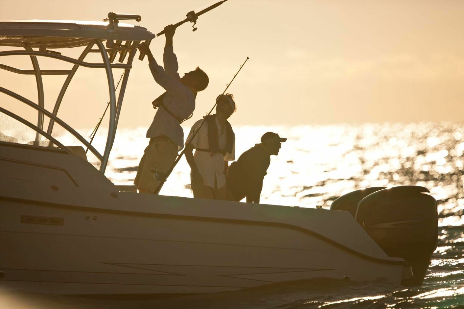 License-free image Three people on a boat at dusk with fishing rods, silhouetted against the shimmering water. One person stands holding up a fishing rod, while the other two stand and sit, looking out towards the sea.