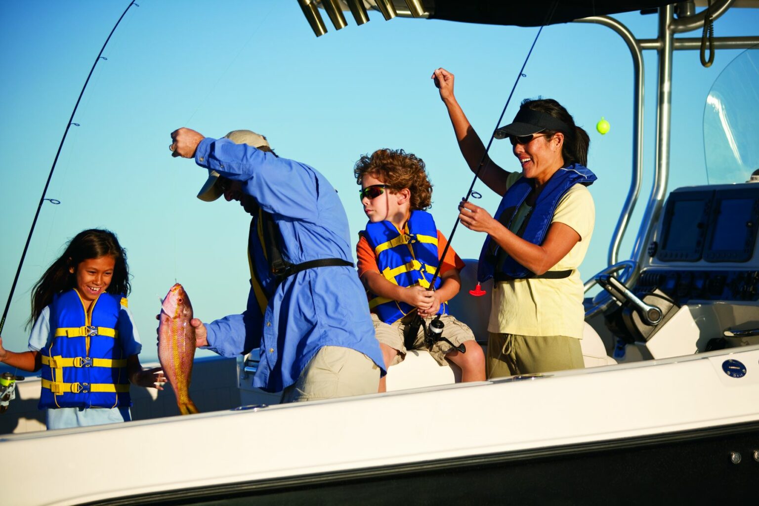 License-free image - A family on a boat, all wearing life jackets, is fishing together. A man is holding up a freshly-caught fish while a young boy and girl look excited. An adult woman stands nearby, smiling and raising a fishing rod in celebration. The sky is clear and sunny.