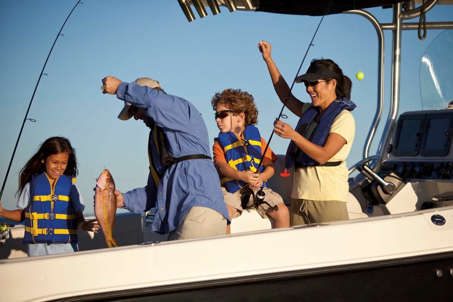 License-free image - A family of four is on a boat, fishing together. An older man, wearing a hat and blue shirt, holds a freshly caught fish. A young boy and girl, both in life jackets, look excited, while a woman in sunglasses and a hat raises her arm, celebrating the catch.