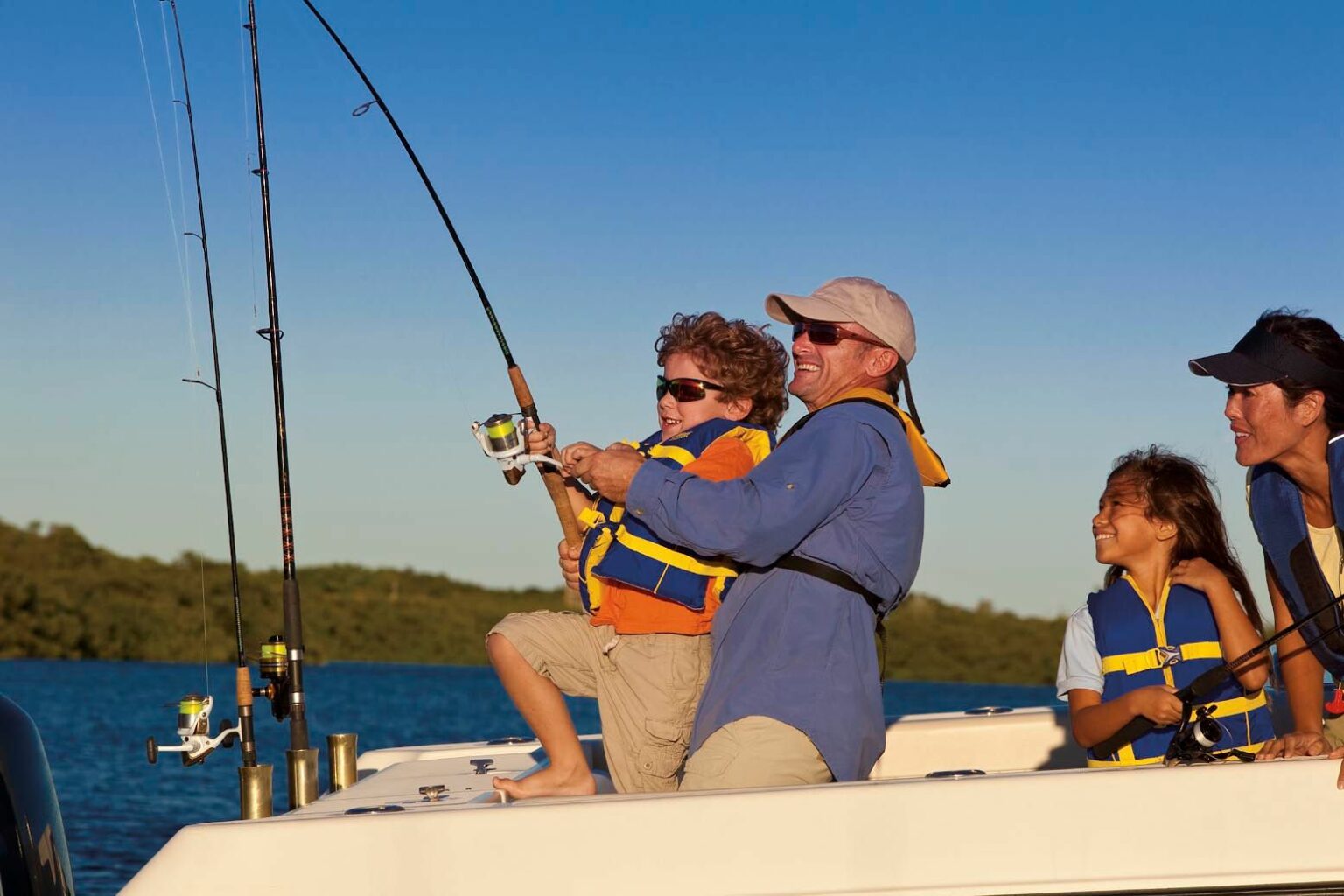 License-free image - A man helps a child reel in a fish aboard a boat, while another adult and child look on. All are wearing life jackets and appear to be enjoying the sunny day on the water. The background shows calm waters and a distant treeline.