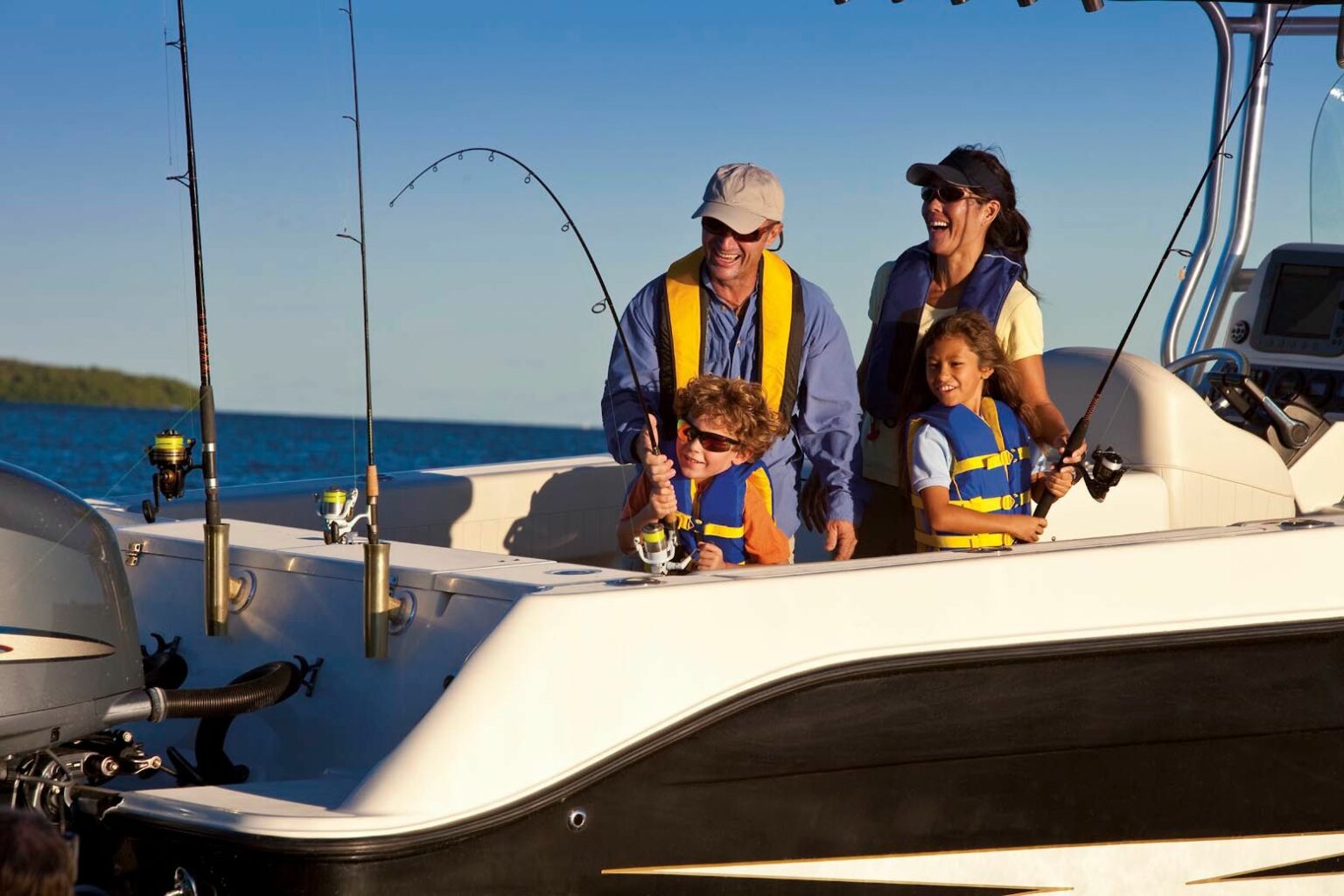 License-free image - A happy family dressed in life jackets enjoys a day of fishing on a boat. Two children are handling fishing rods with the help of their parents, who stand behind them, smiling. The sun is shining, and the boat is surrounded by a serene body of water.