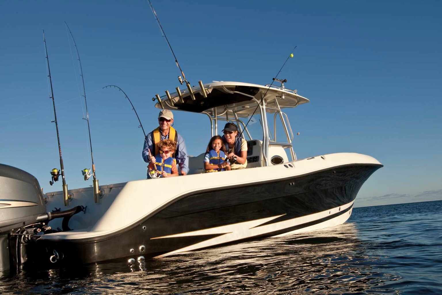 License-free image. Three people enjoy their time on a sleek, white fishing boat out on the water under a clear blue sky. The people, wearing life jackets and sitting at the edge of the boat, appear to be engaged in fishing, as multiple fishing rods are mounted at the stern.