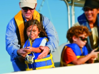 License-free image. An adult in sunglasses helps a young girl hold a fishing rod while another child in sunglasses looks on. All are wearing life jackets, suggesting they are on a boat. A fourth person smiles in the background.