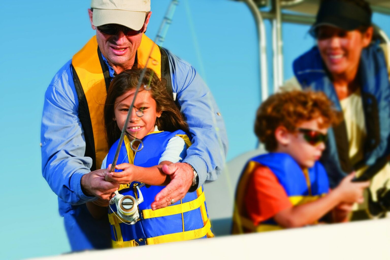 License-free image. An adult in sunglasses helps a young girl hold a fishing rod while another child in sunglasses looks on. All are wearing life jackets, suggesting they are on a boat. A fourth person smiles in the background.