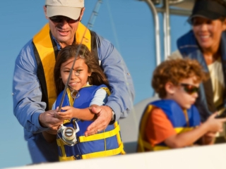 Royalty-free image A man with a white cap helps a smiling young girl in a yellow life jacket hold a fishing rod on a boat. A woman and a boy, also in life jackets, are in the background, engaged in their activities. All seem to be enjoying a sunny day out on the water.