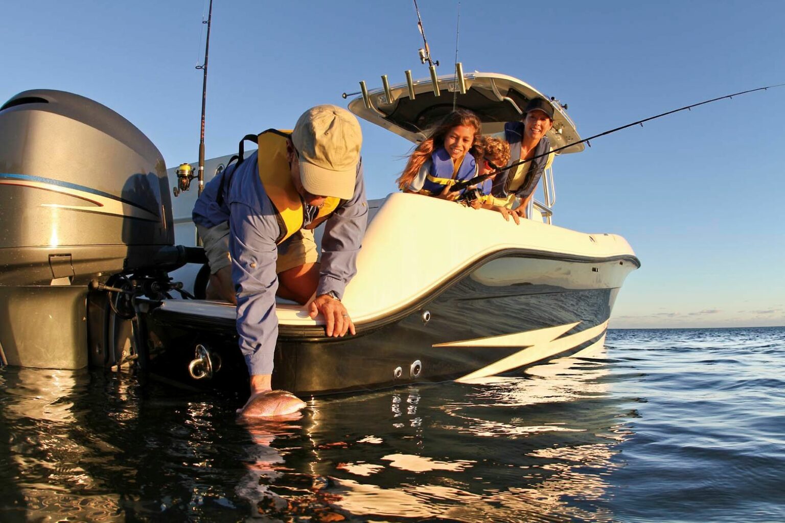 Royalty-free image. Three people wearing life vests are on a boat in calm waters. One person leans over the side, gently releasing a fish back into the water. The other two are smiling and sitting in the boat. It&#039;s a sunny day, and they appear to be enjoying a fishing outing.