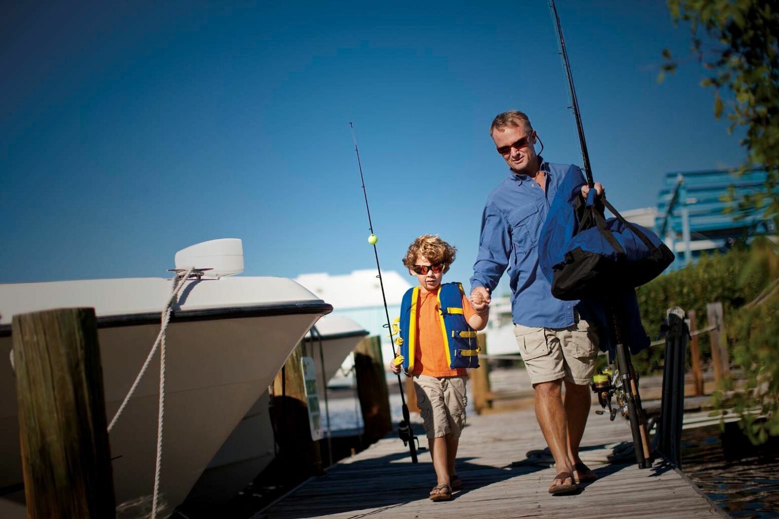 License-free image - A man and a child walk hand-in-hand on a sunny day along a wooden dock, each holding a fishing rod. The child sports a life jacket and sunglasses, while the man carries a black bag. In the background, a boat is moored beside the dock, and the sky is clear and blue.