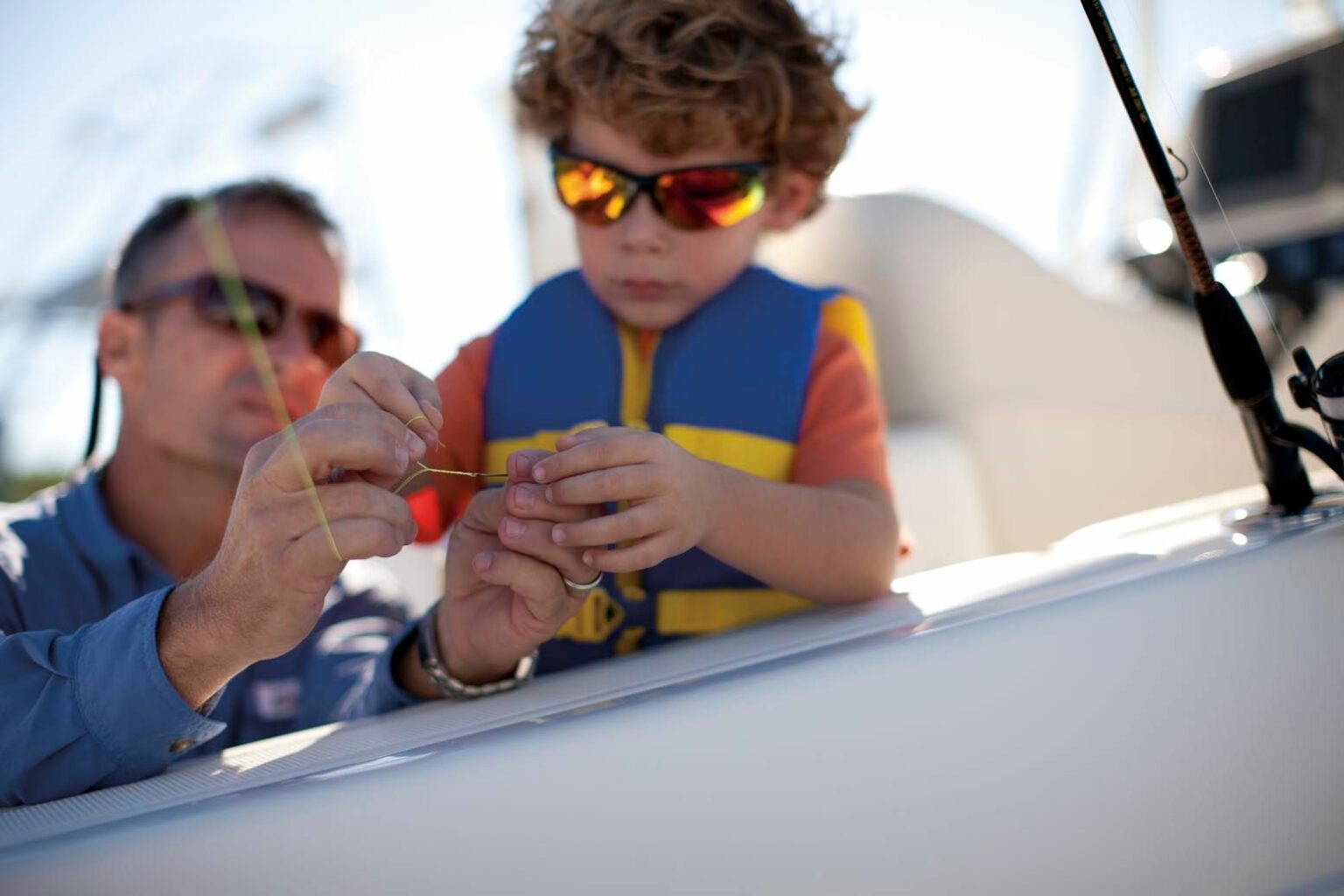 License-free image A man and a young child wearing sunglasses and a life vest are focused on a small object held between the man’s fingers. They appear to be on a boat. The background is blurry, with indistinct poles and equipment visible.