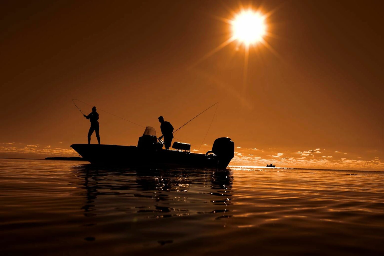 License-free image - Silhouetted image of two people fishing from a small boat on calm water during sunrise or sunset. The sky is a warm orange hue, and the sun shines brightly above the horizon, casting reflections on the water&#039;s surface.