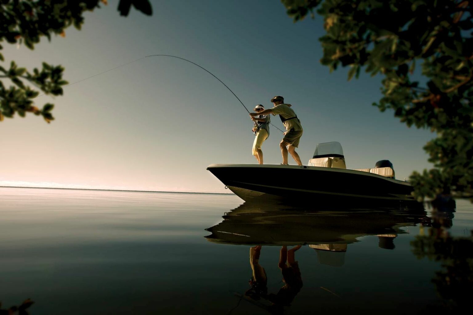 License-free image - Two people stand on a boat silhouetted against a sunset, fishing on calm waters. One person is reeling in a fish while the other assists. The scene is framed by leafy branches on both sides, and the water reflects the tranquility of the moment.