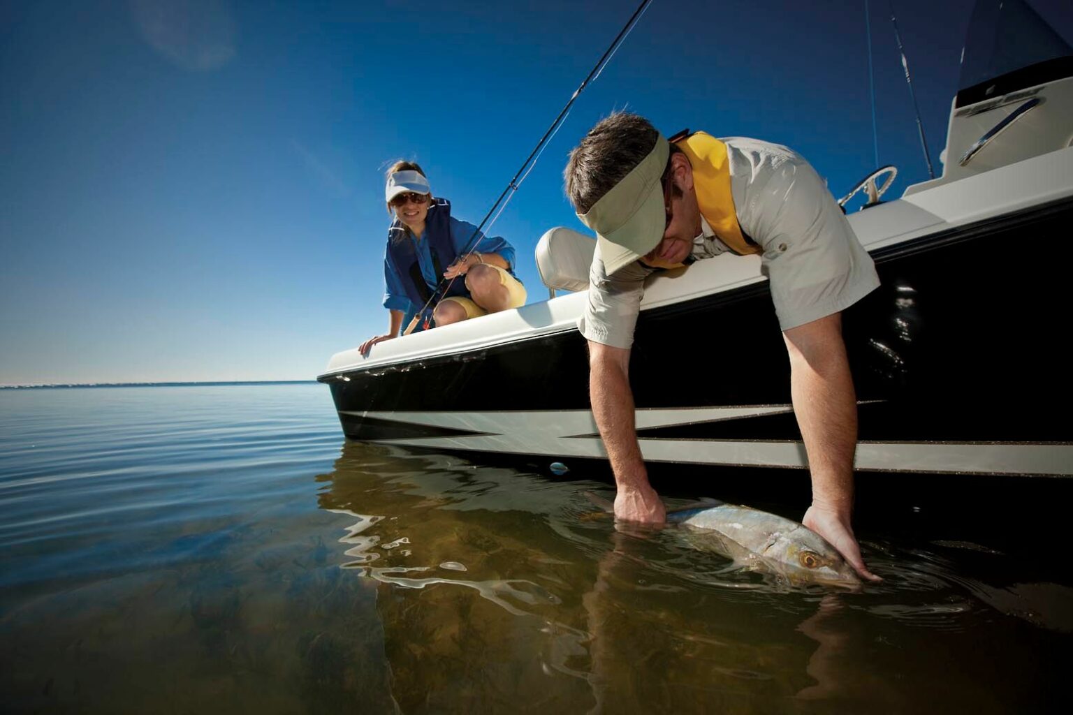 License-free image. A man wearing a beige cap and white shirt releases a fish back into the water from a boat while a woman wearing a blue shirt and white cap watches. The scene is set on a calm, clear day with blue skies and tranquil waters.