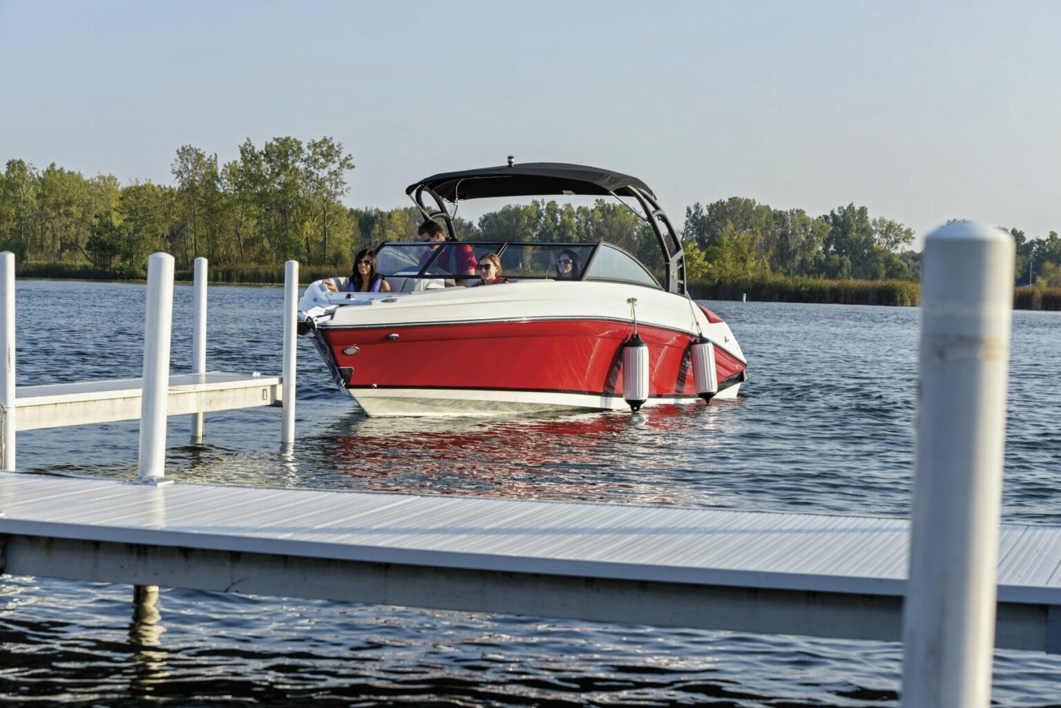 License-free image. A red and white motorboat with several people on board is approaching a dock on a calm body of water. The dock is partially visible in the foreground, and the background consists of lush greenery and trees under a clear sky.