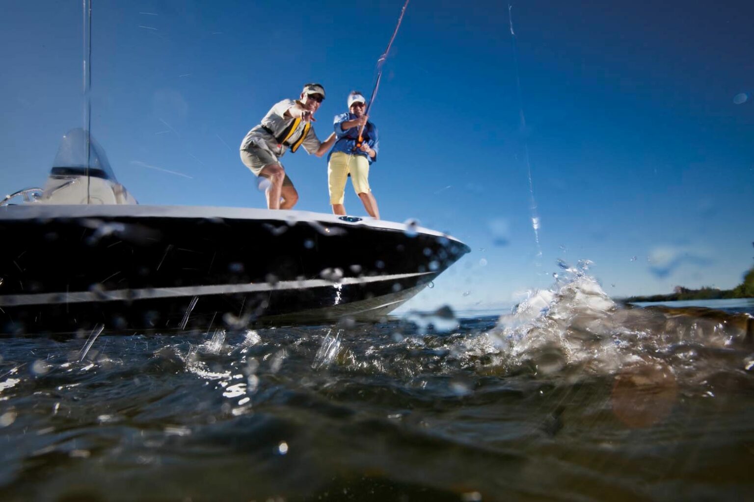 License-free image Two people stand on the deck of a boat, actively fishing. One person leans over the railing, seemingly reeling in a catch, while the other stands behind them. Water splashes near the boat, and the sky above is clear and blue.