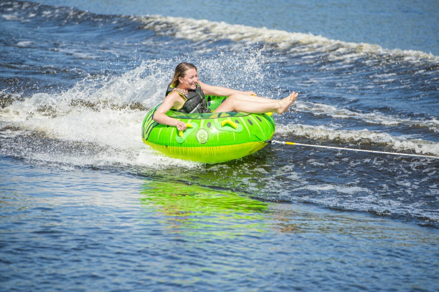 License-free image - A person wearing a life jacket is joyfully riding an inflatable green tube being towed across the water. They are splashing through waves, with a big smile on their face and feet kicked up in the air.